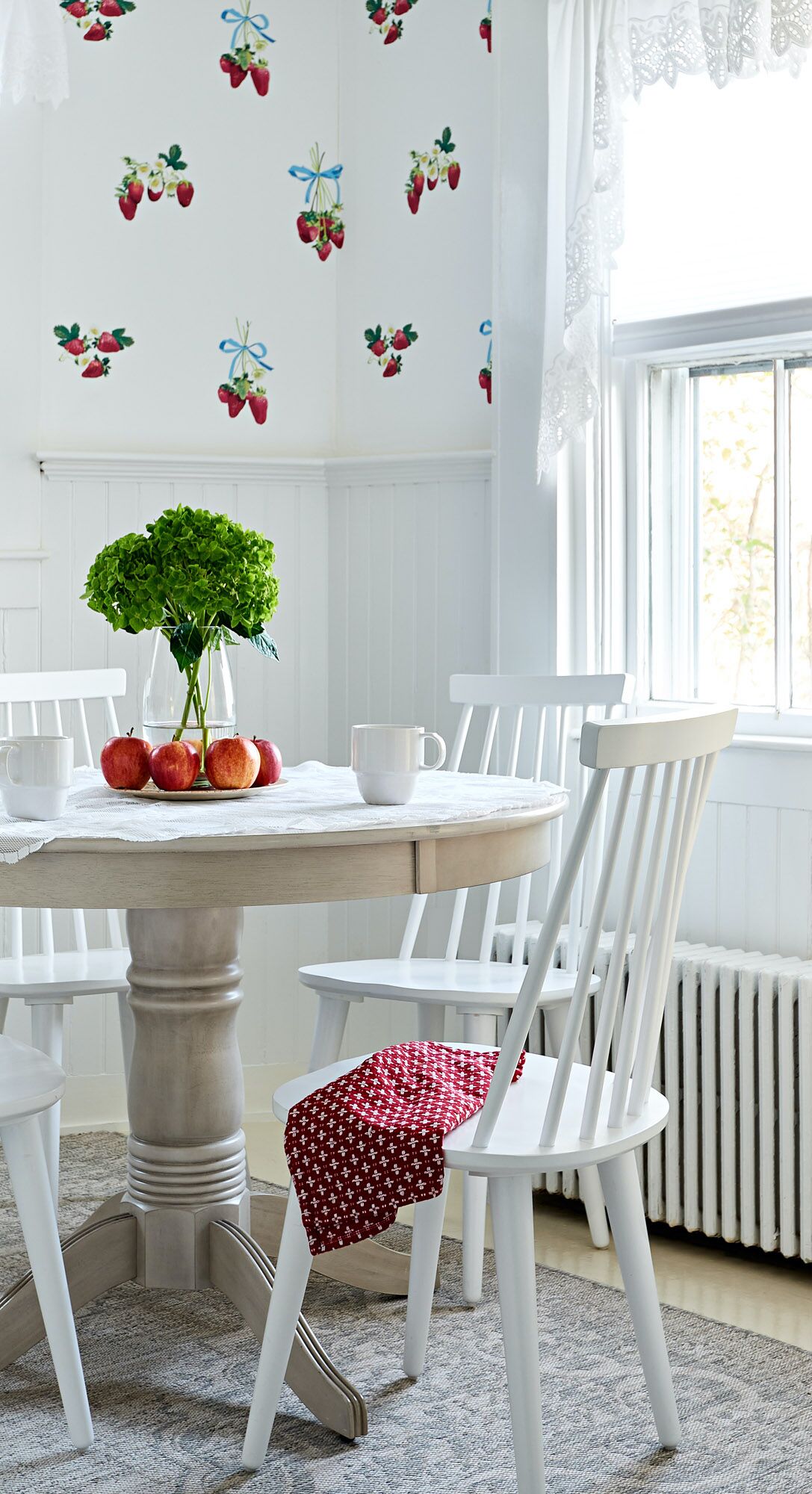 Un comedor con una mesa redonda, sillas blancas y un pared decorada con motivos de fresas.