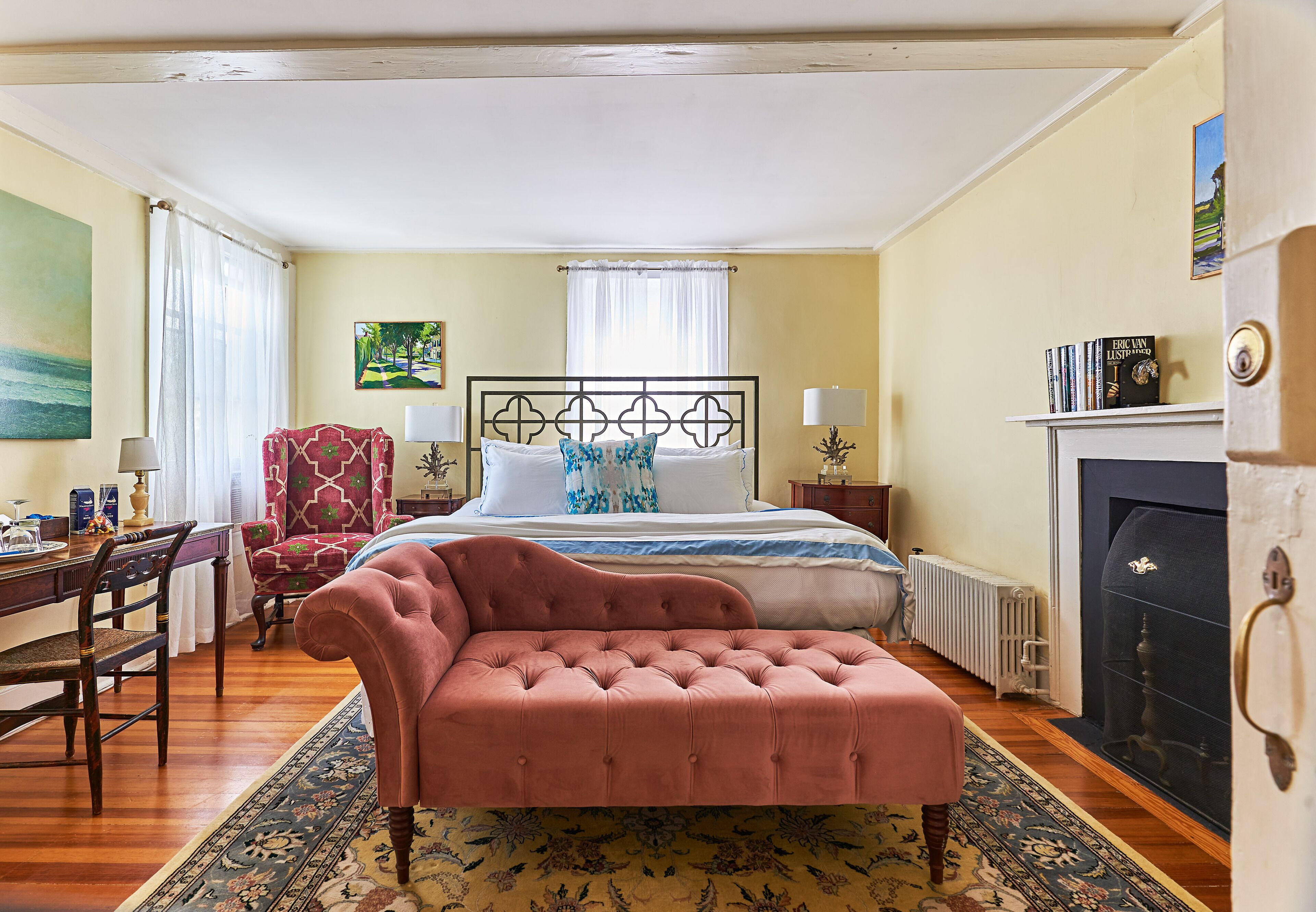 A bedroom with a large bed, a sitting area with a chair, a desk, and a fireplace.
