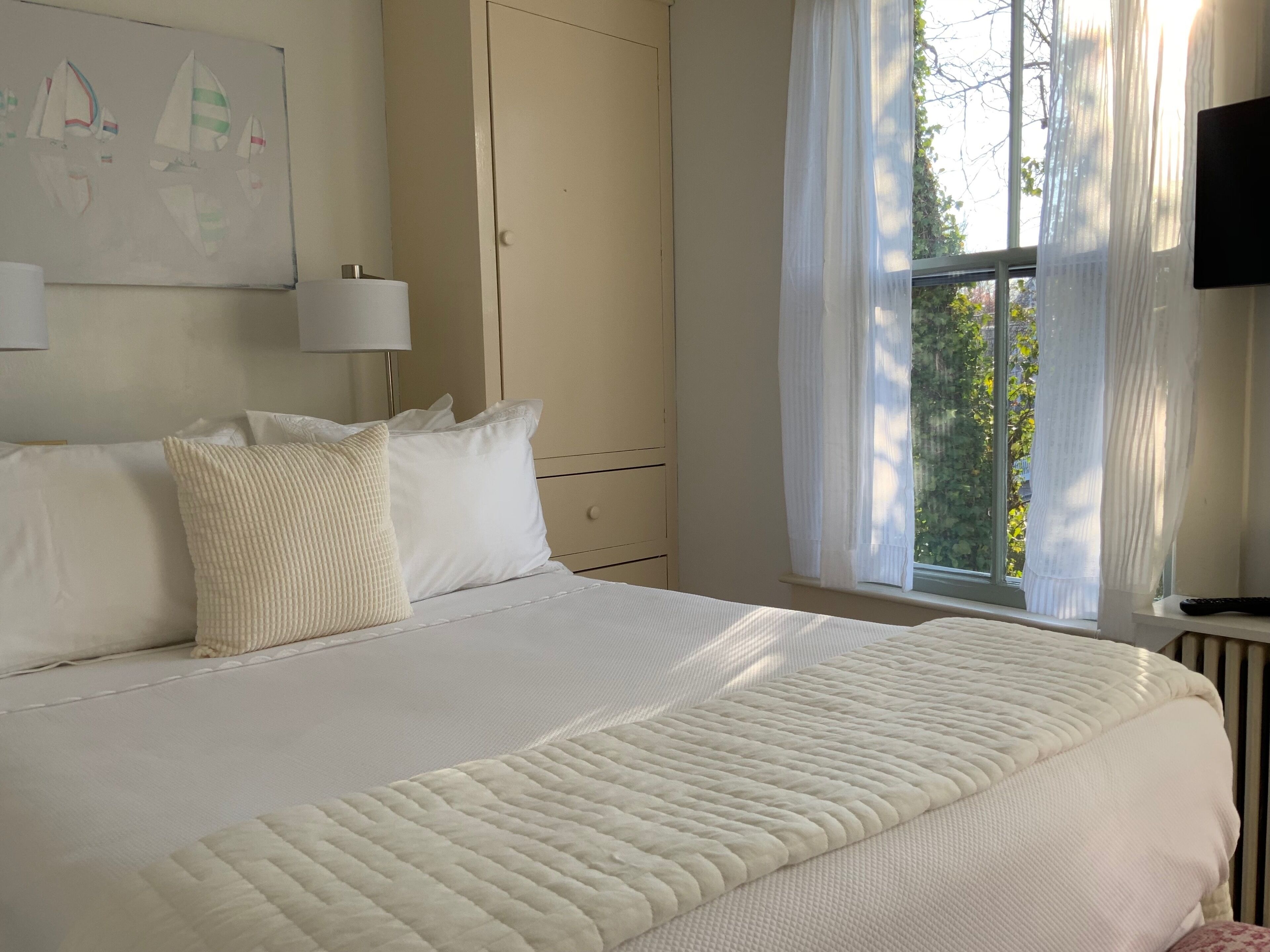 Une chambre à coucher avec un grand lit, une fenêtre avec des rideaux, un tableau accroché au mur et un téléviseur posé sur un meuble.