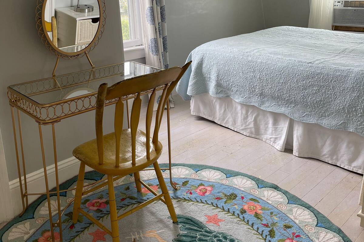 A bedroom with a bed, a wooden chair, a small table with a mirror, and a decorative rug.