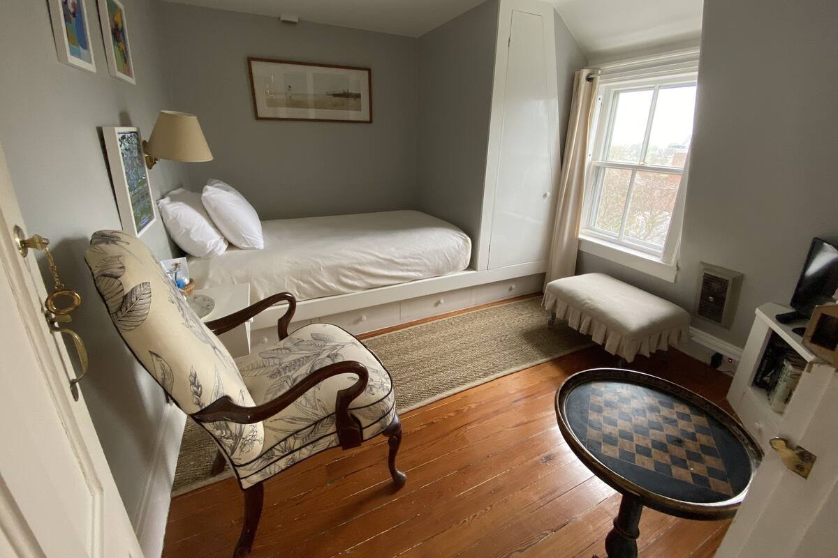 A bedroom with a single bed, a chair, a small table, a window, and a picture on the wall.