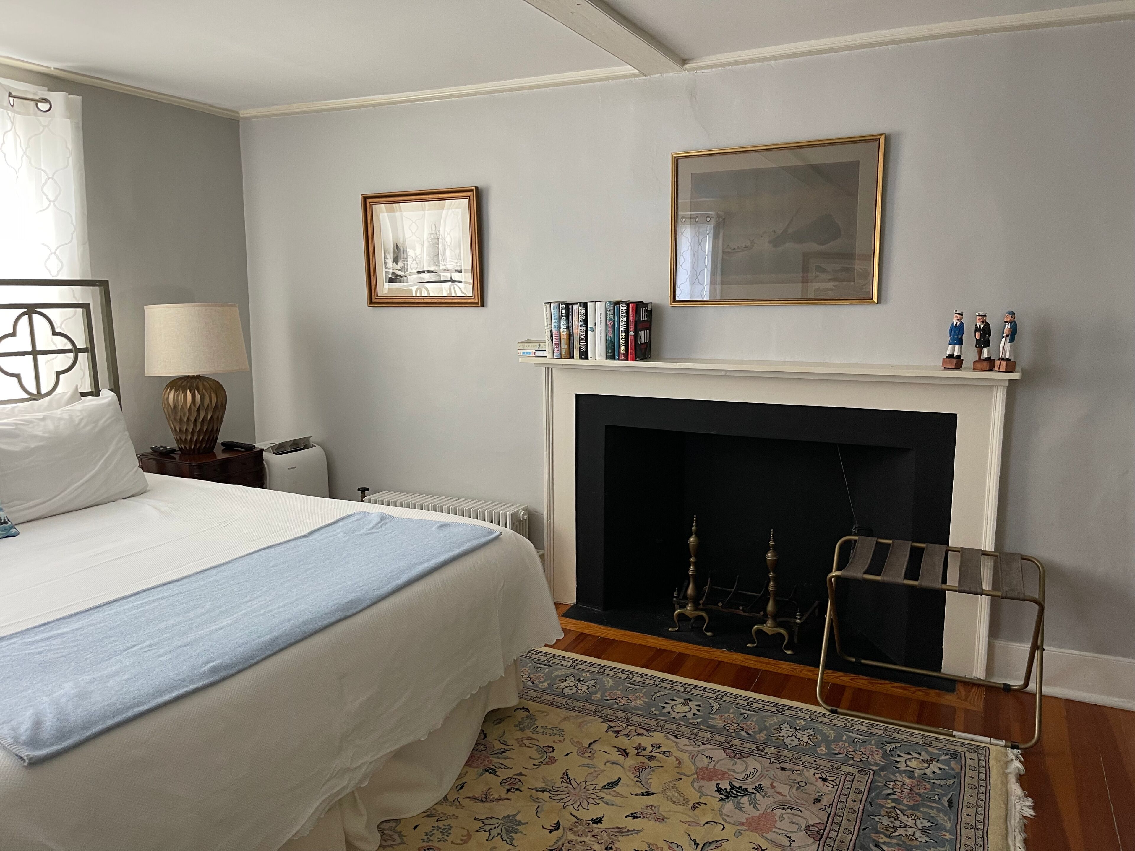 A bedroom with a bed, a nightstand, a lamp, a fireplace, a rug, and a framed picture on the wall.