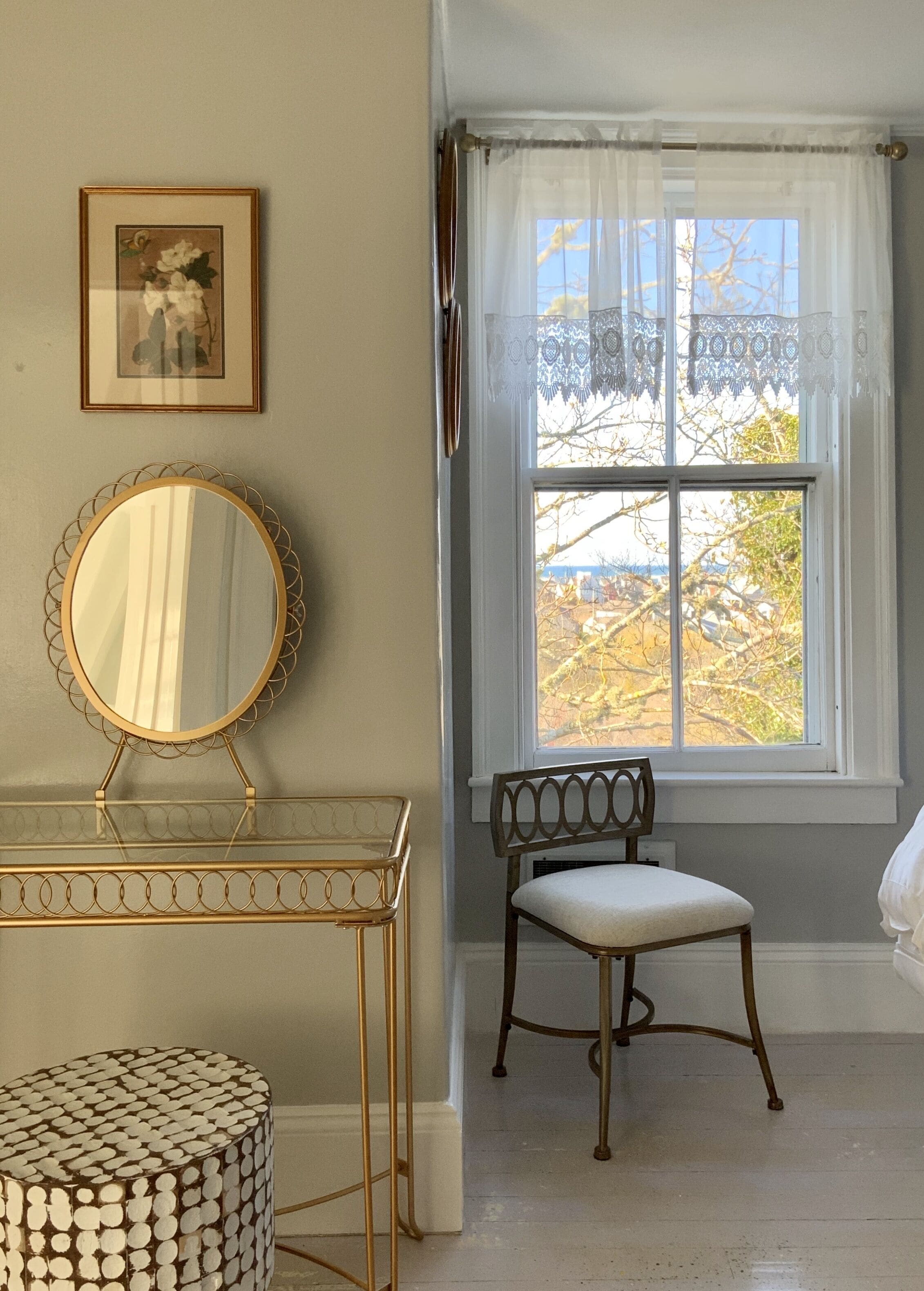 A room with a round mirror on a gold-framed table, a chair with a cushioned seat, and a window with lace curtains.