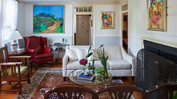 A living room with a sofa, armchairs, a coffee table with books and plants, a fireplace, and two paintings on the wall.