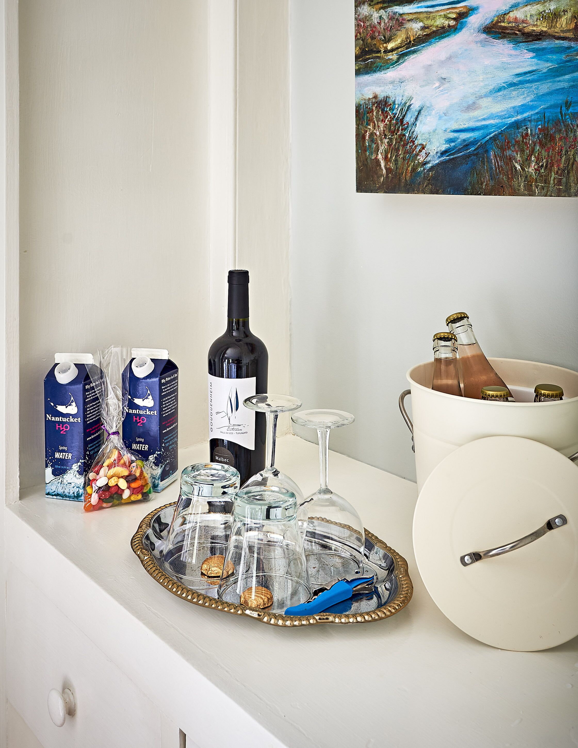 A tray with a bottle of wine, glasses, a bucket, and water cartons on a white shelf.