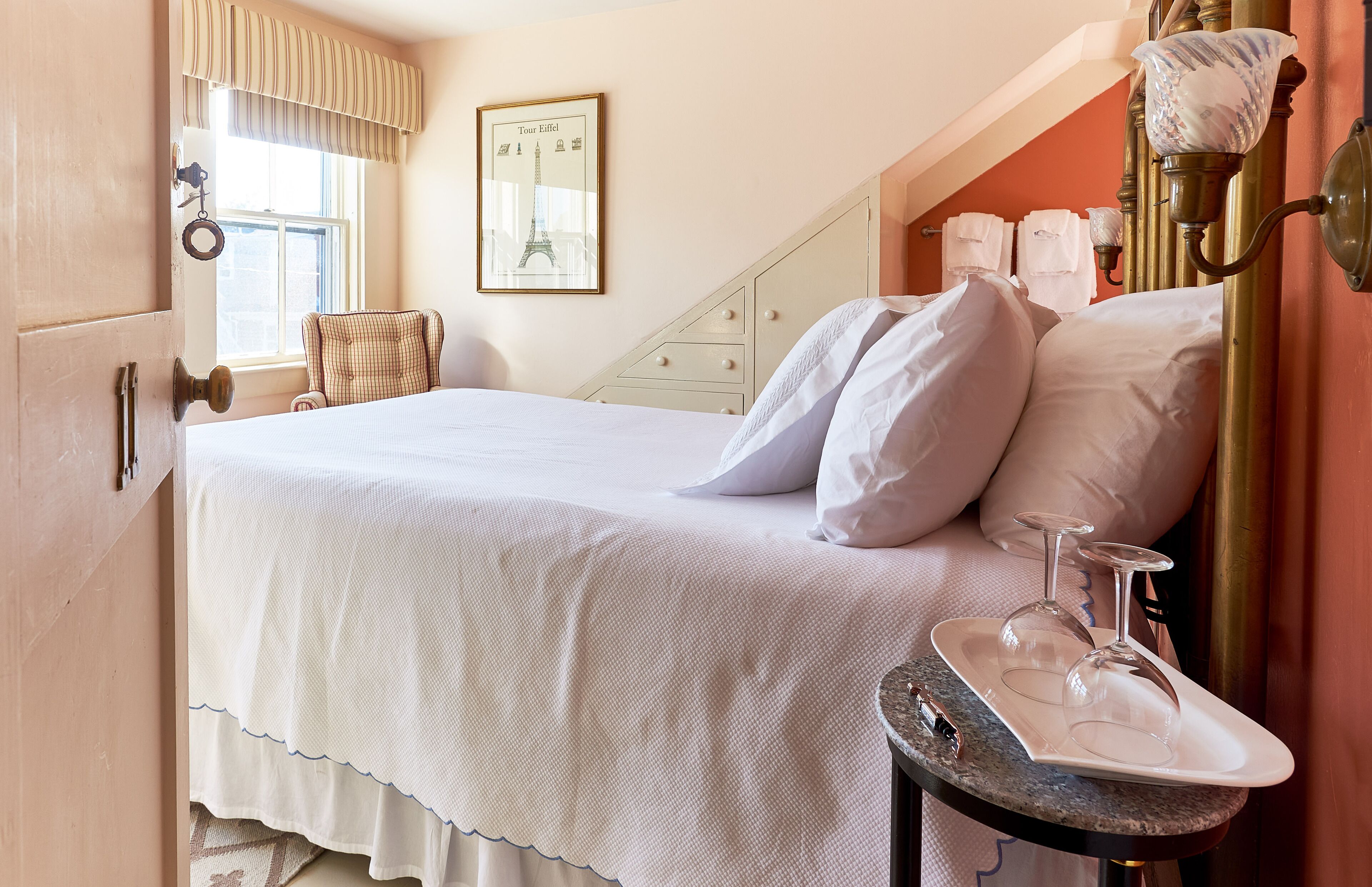 A neatly made bed with white linens, a bedside table with a tray holding wine glasses, and a framed picture of the Eiffel Tower on the wall.