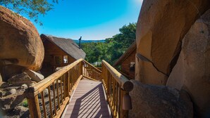 A wooden bridge leading to a log cabin with a thatched roof, surrounded by large rocks and trees.