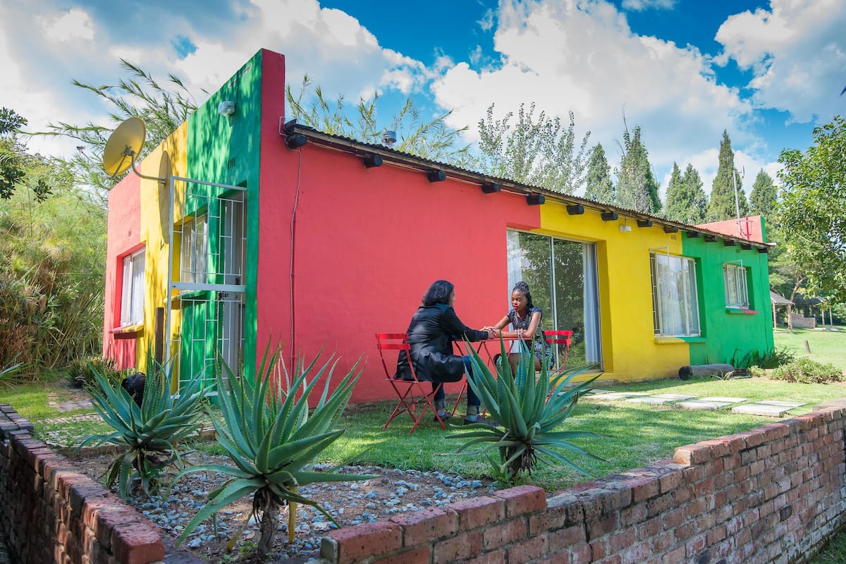 A colorful house with a red, yellow, and green facade, a satellite dish, and two people sitting at a table outside.