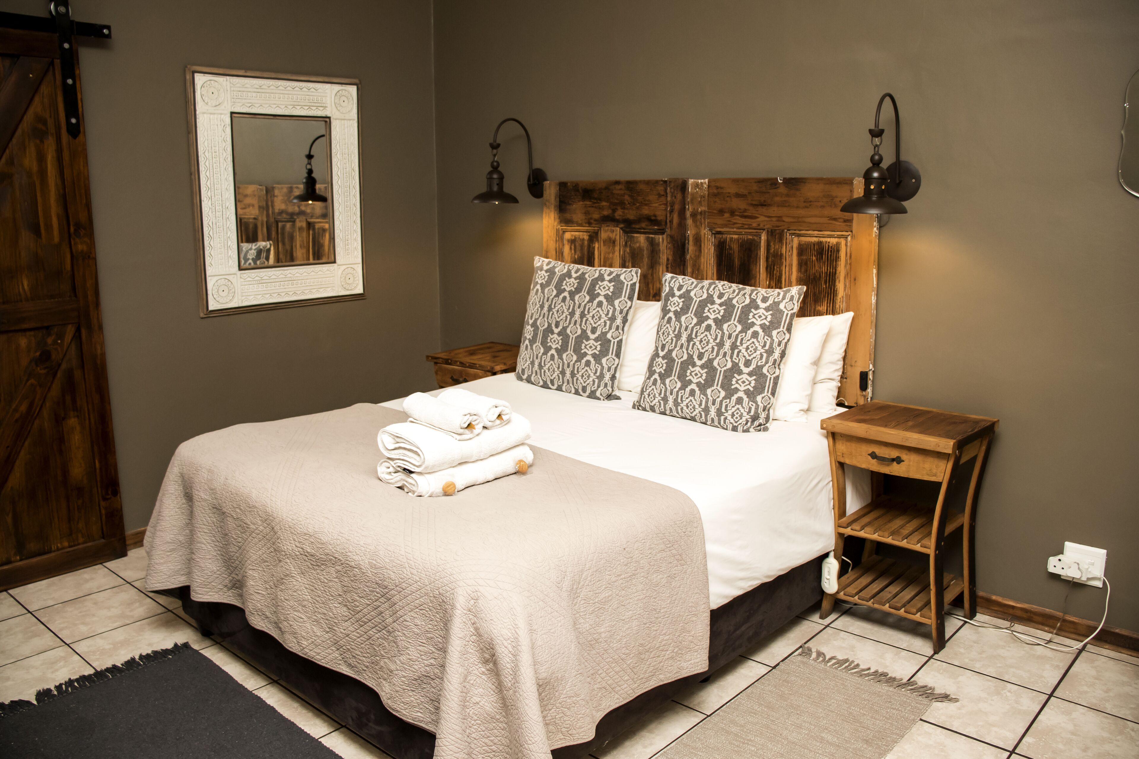 A neatly made bed with a beige quilt, a wooden headboard, and patterned pillows. There is a small wooden nightstand with a lamp and a power outlet. A black rug is on the floor, and a wooden door is visible to the left.