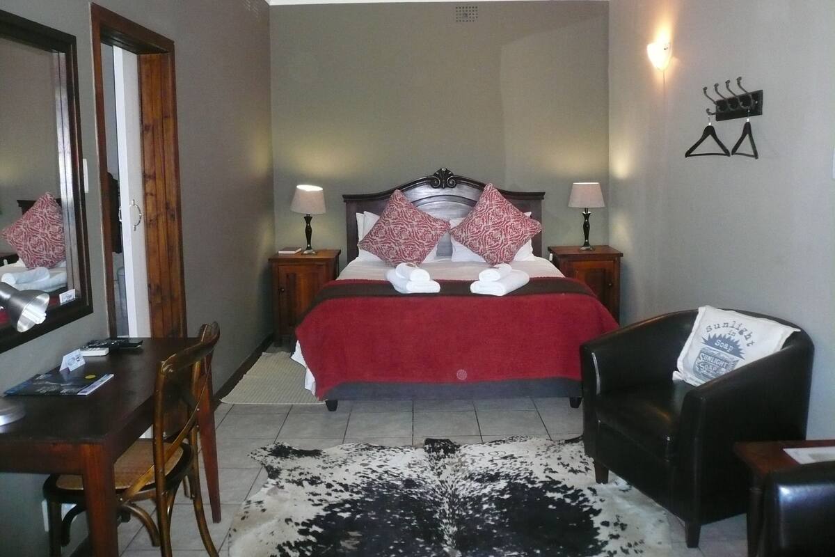 Ein Hotelzimmer mit Bett, Nachttischlampen, Schreibtisch, Stuhl, Spiegel und einem Kuhfellteppich.