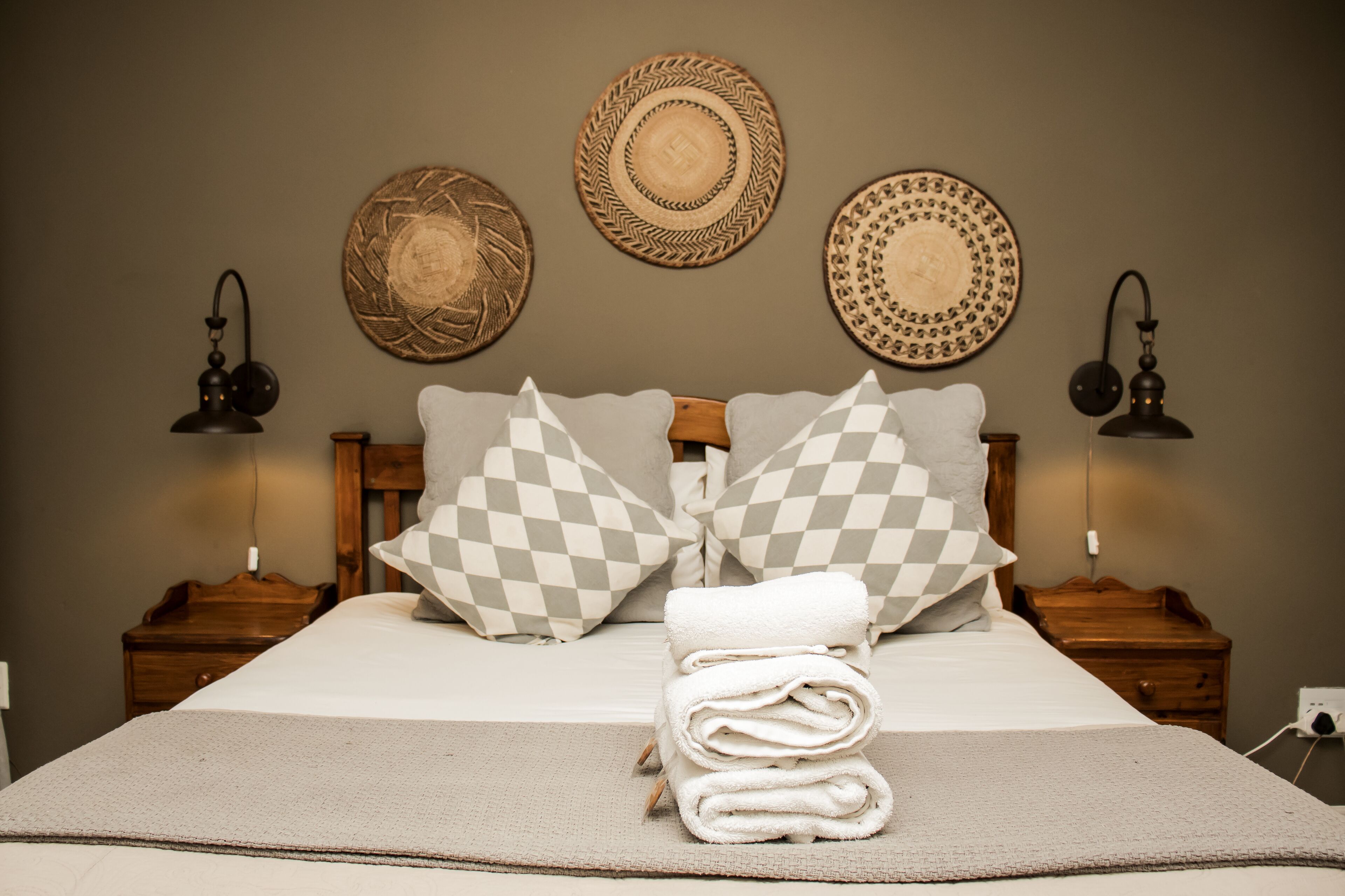 A neatly made bed with checkered pillows, a folded towel, and decorative woven plates on the wall.