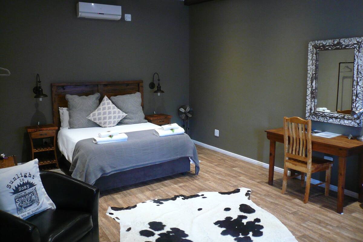 Ein Schlafzimmer mit einem Bett, einem Schreibtisch, einem Stuhl, einem Spiegel, einer Lampe und einem Kuhfellteppich.