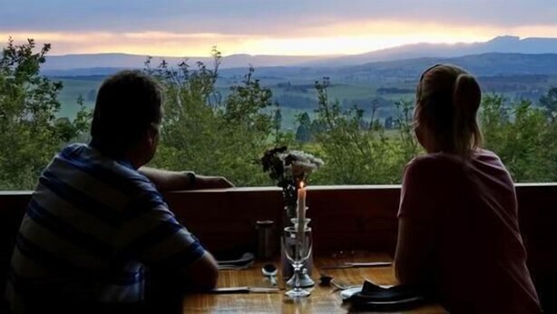 Twee personen zitten aan een tafel met een kaars, met uitzicht op een prachtig landschap.