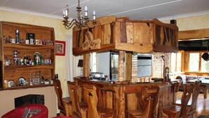 Een rustieke bar met houten toonbank, voorzien van een inbouwkoelkast, een kroonluchter en een zithoek met houten stoelen en een rode bank.