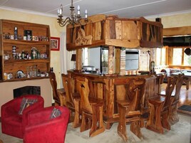 Una zona rústica de bar con barra de madera, refrigerador empotrado, una lámpara de araña y un espacio de asientos con sillas de madera y un sofá rojo.