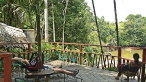 Terraza de madera al aire libre con sillas y mesas de metal, rodeada por una barandilla de madera, con vistas a un bosque frondoso y verde.