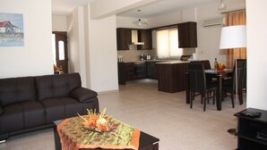 A modern living room with a dark brown sofa, a dining table with chairs, and a kitchen area with dark wooden cabinets and a central island. - Vineland Holiday Villas (Pissouri)