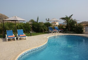 A swimming pool with lounge chairs and umbrellas, surrounded by greenery and a clear sky. - Vineland Bay Villas (Pissouri)