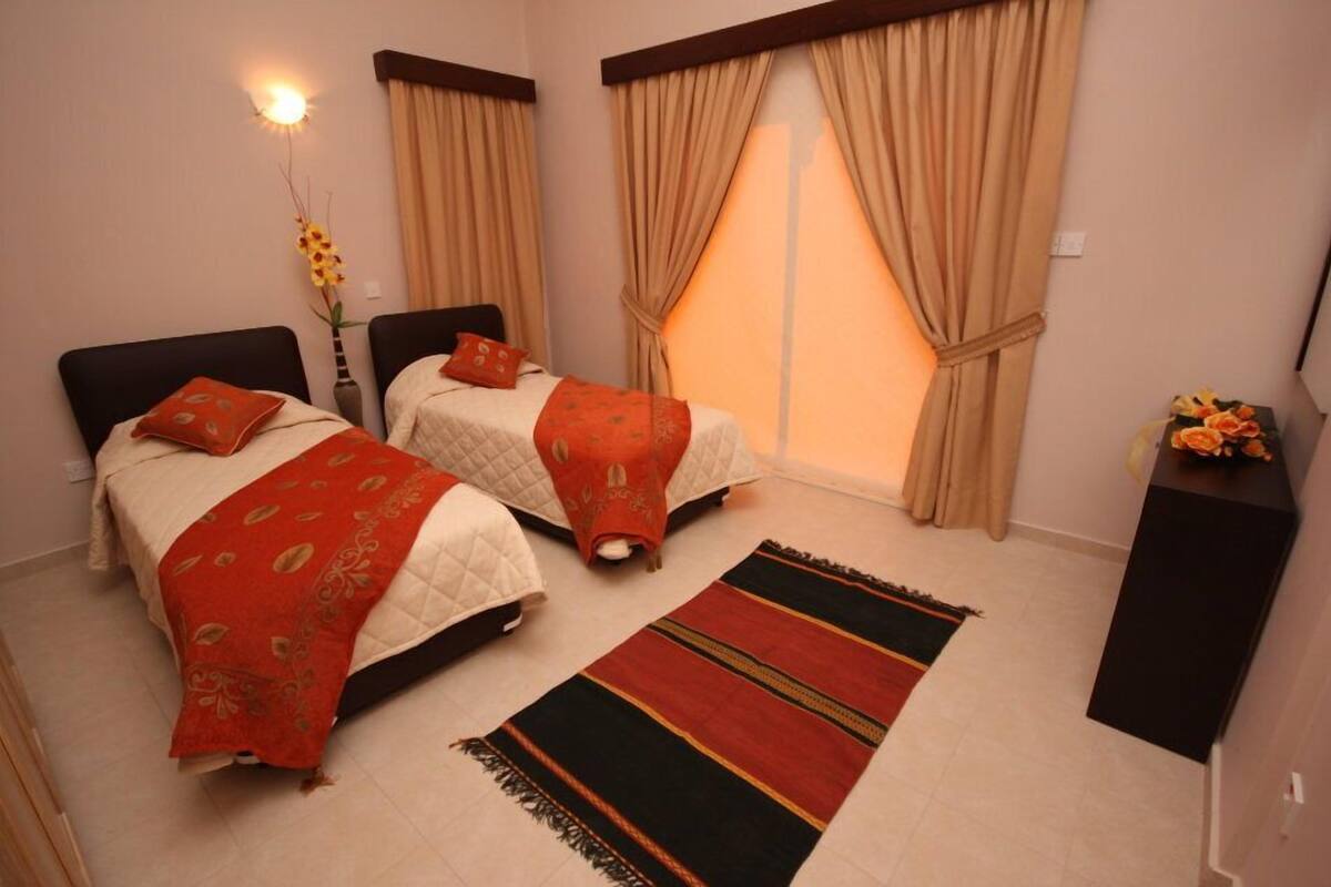 Un dormitorio con dos camas, cortinas color crema, una mesita de noche, un jarrón con flores y una alfombra a rayas.