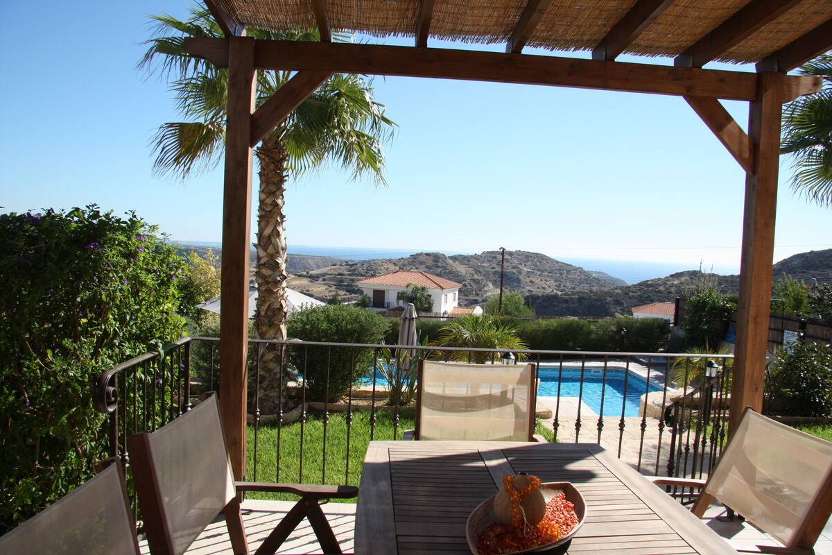 Terraza de madera con mesa y sillas, con vista a una piscina y un paisaje pintoresco de colinas y un pueblo lejano.