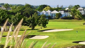 Een golfbaan met bunkers en een vlaggenstok, omgeven door weelderig groen en witte gebouwen.