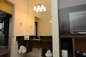 A bathroom with a dark wooden vanity, a mirror, and a sink. There are towels stored under the vanity and a toilet paper roll visible. - Premiere Suites - St. John's Signal Hill (St. John's)