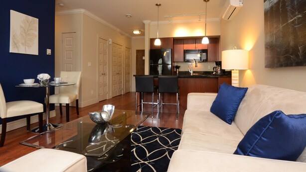A modern living room with a glass coffee table, a beige sofa with blue cushions, a dining area with a small table and chairs, and a kitchen area with a refrigerator, microwave, and bar stools.