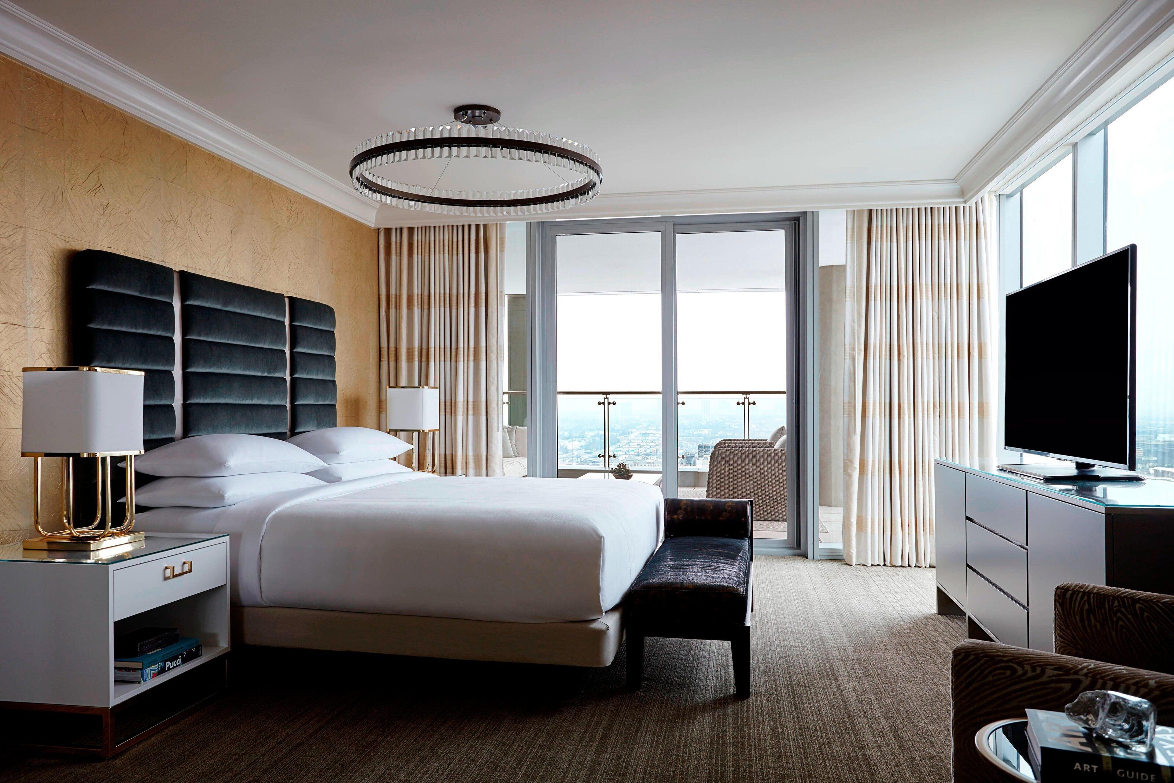 A modern hotel room with a large bed, a desk with a flat-screen TV, a bench, and a view of the city through large windows.