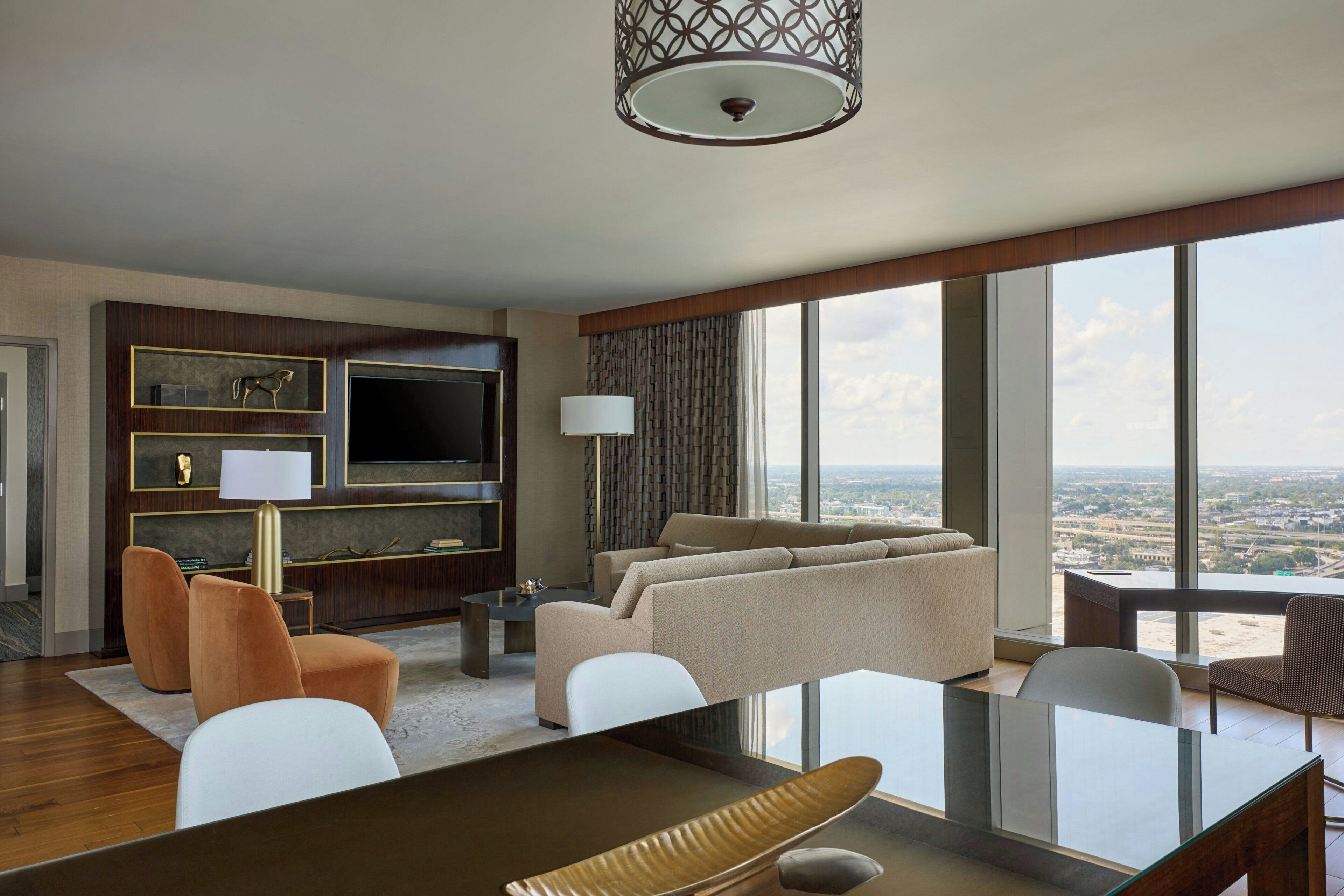 A modern hotel room with a large flat-screen TV, a sofa, a dining table, and a view of the city.