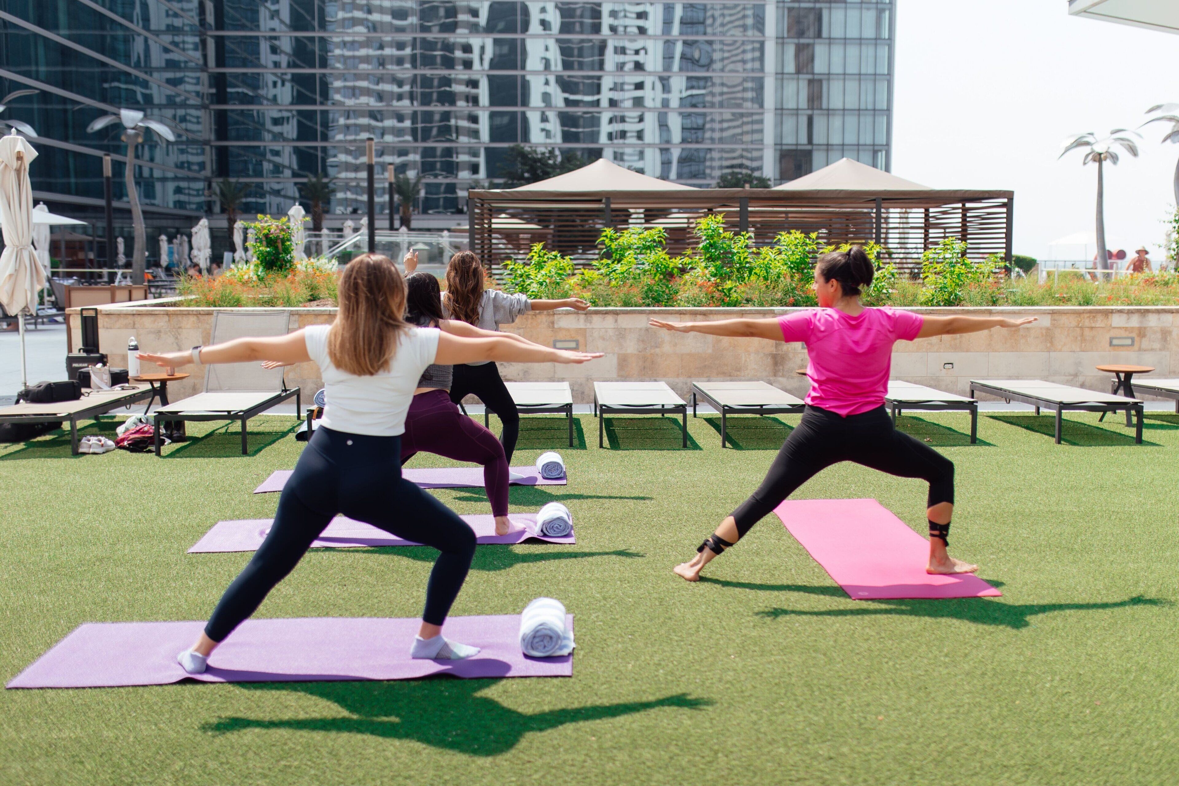 A group of people practicing yoga outdoors on a grassy area with a modern building in the background.