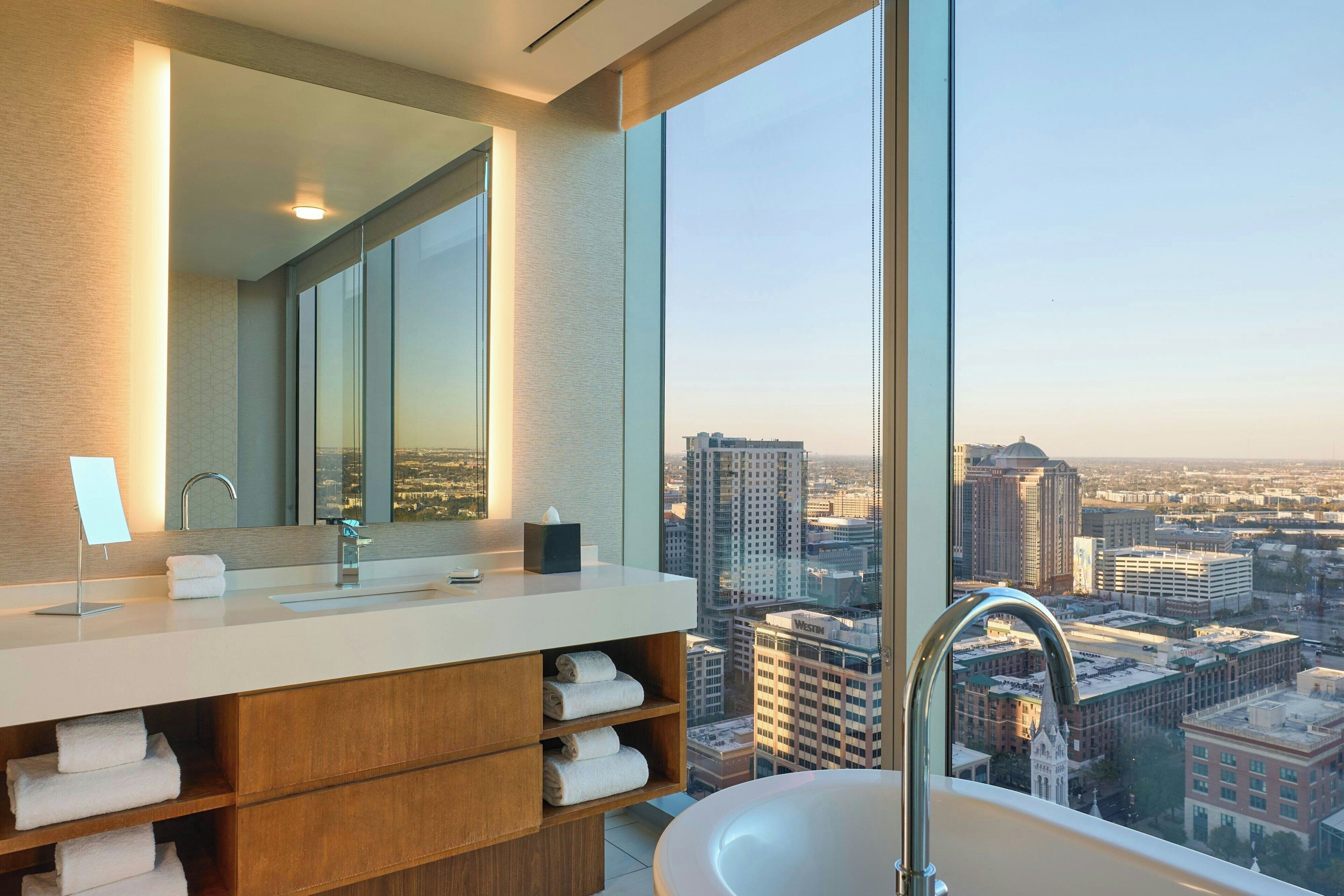 A modern bathroom with a large window, a white sink, a mirror, and a bathtub.