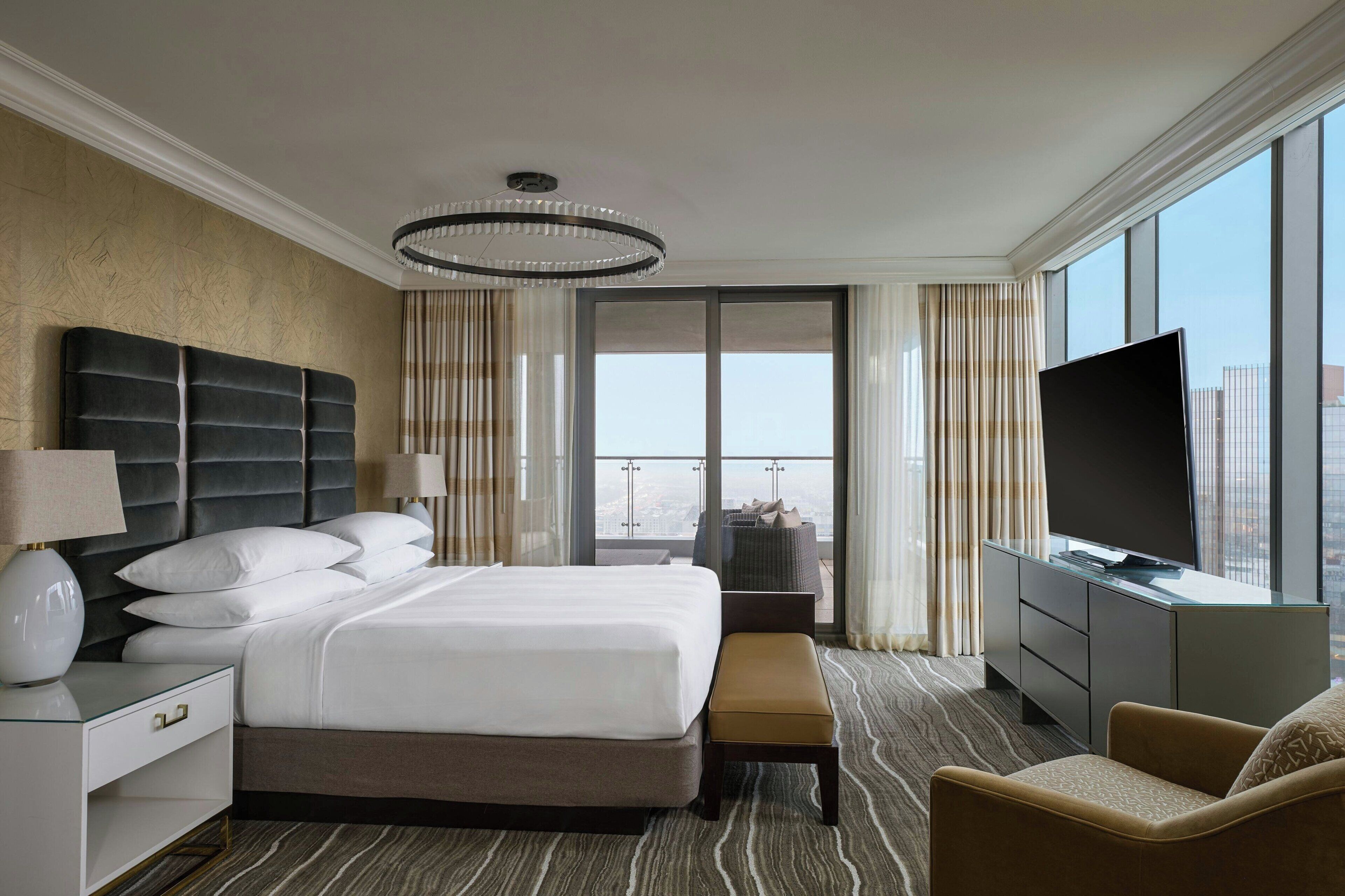 A hotel room with a large bed, a desk with a TV, a chair, and a view of the city through large windows.