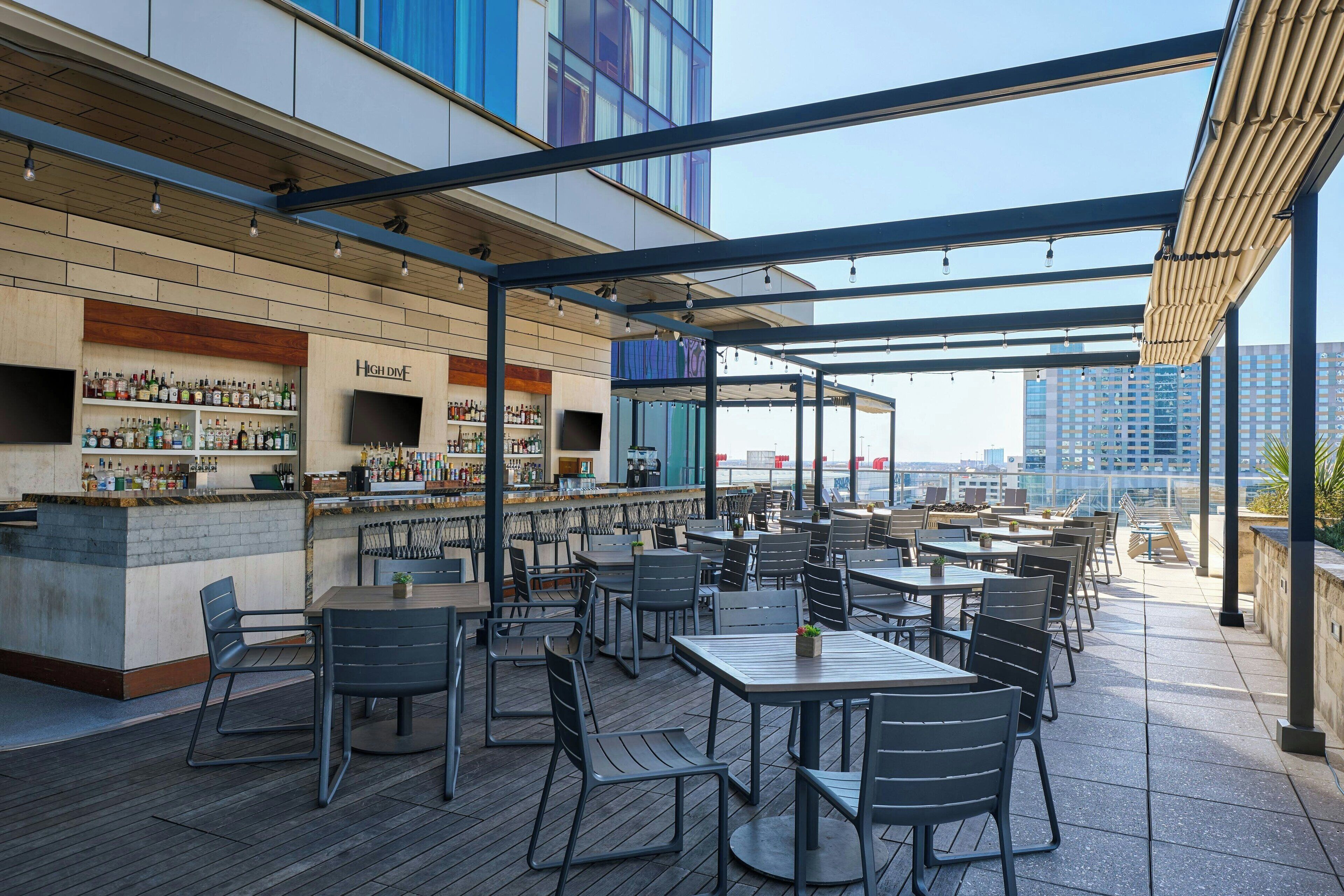 An outdoor dining area with tables and chairs, a bar with shelves of bottles, and a view of the city skyline.