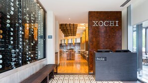 A hotel reception area with a wooden reception desk labeled 'XOCHI' and a 'hola' sign, a bench, and a wall of wine bottles.