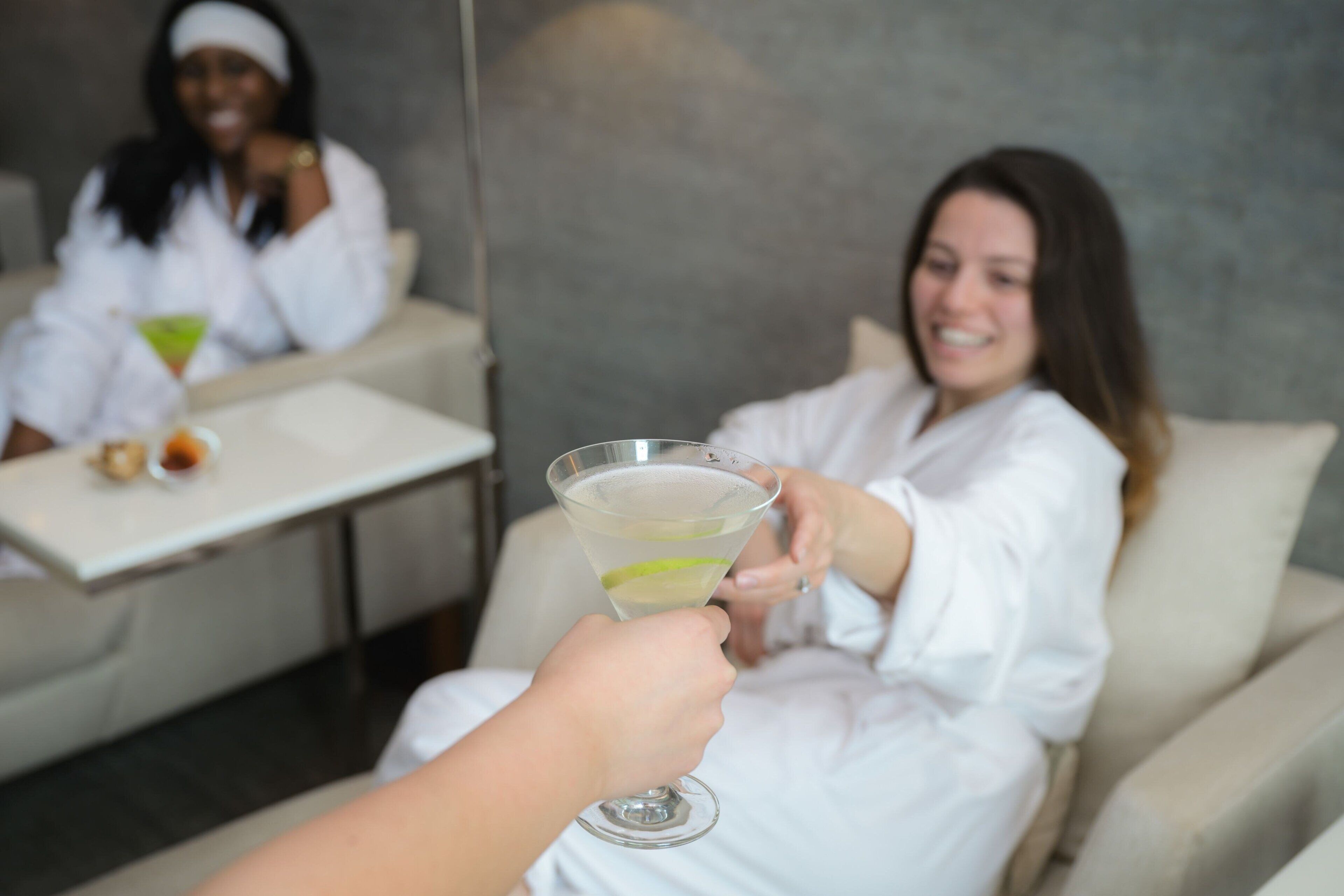 A person holding a martini glass with a lime wedge, facing another person seated at a table with a martini glass.