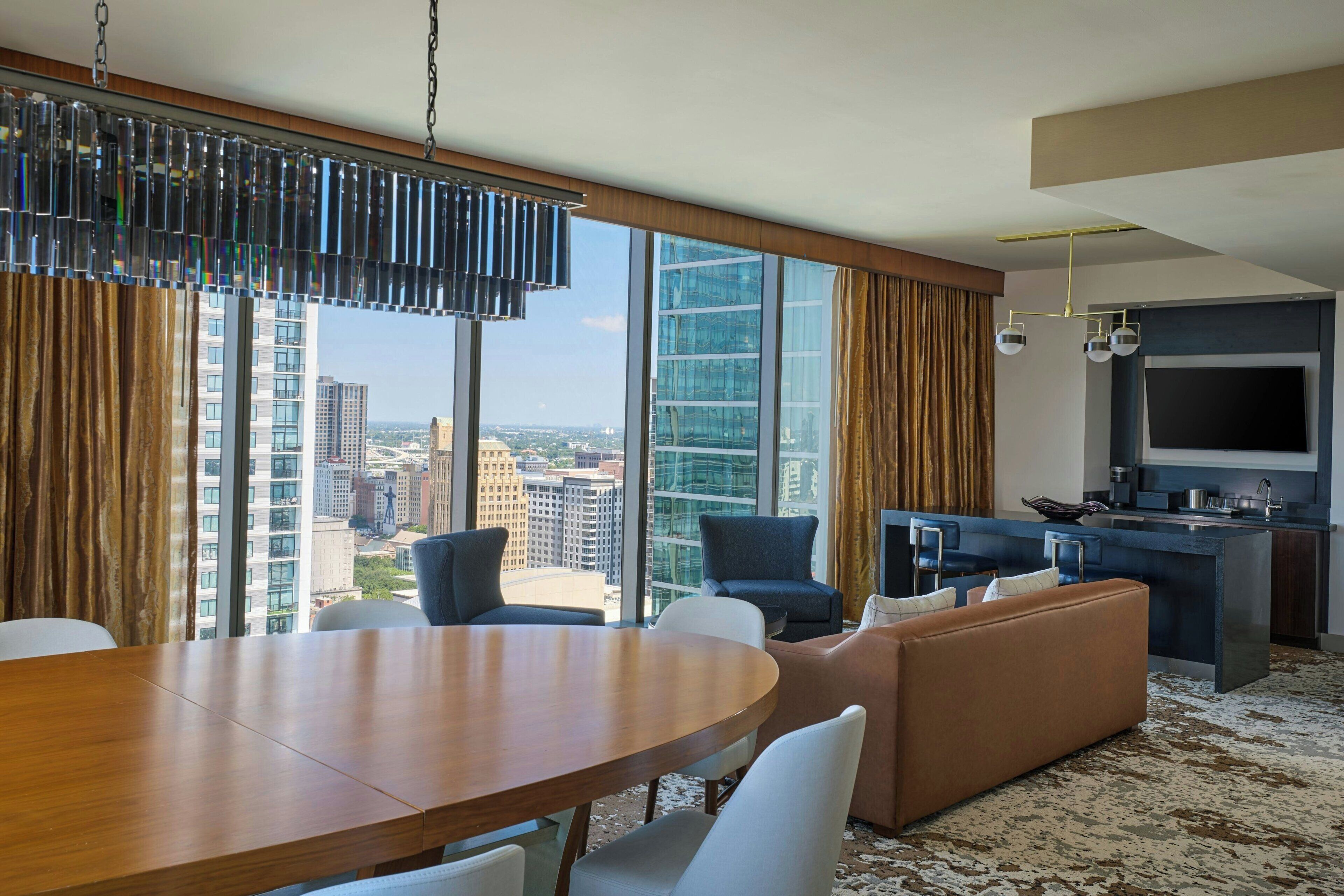 Quarto de hotel moderno com área de refeições, assentos, cozinha e uma grande janela com vista para a cidade.