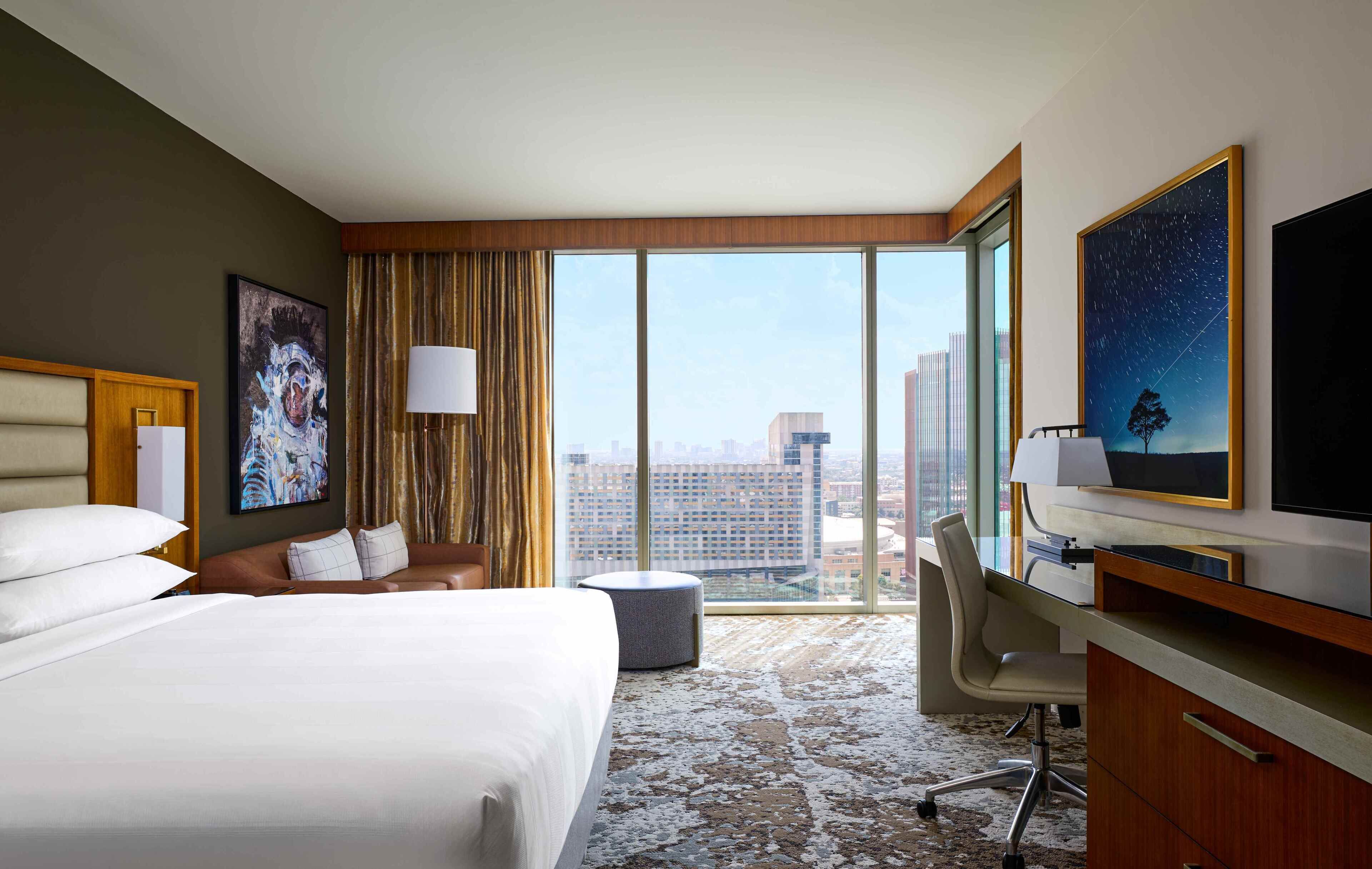 Quarto de hotel com uma cama grande, uma escrivaninha com cadeira, televisão e uma janela ampla com vista para a cidade.