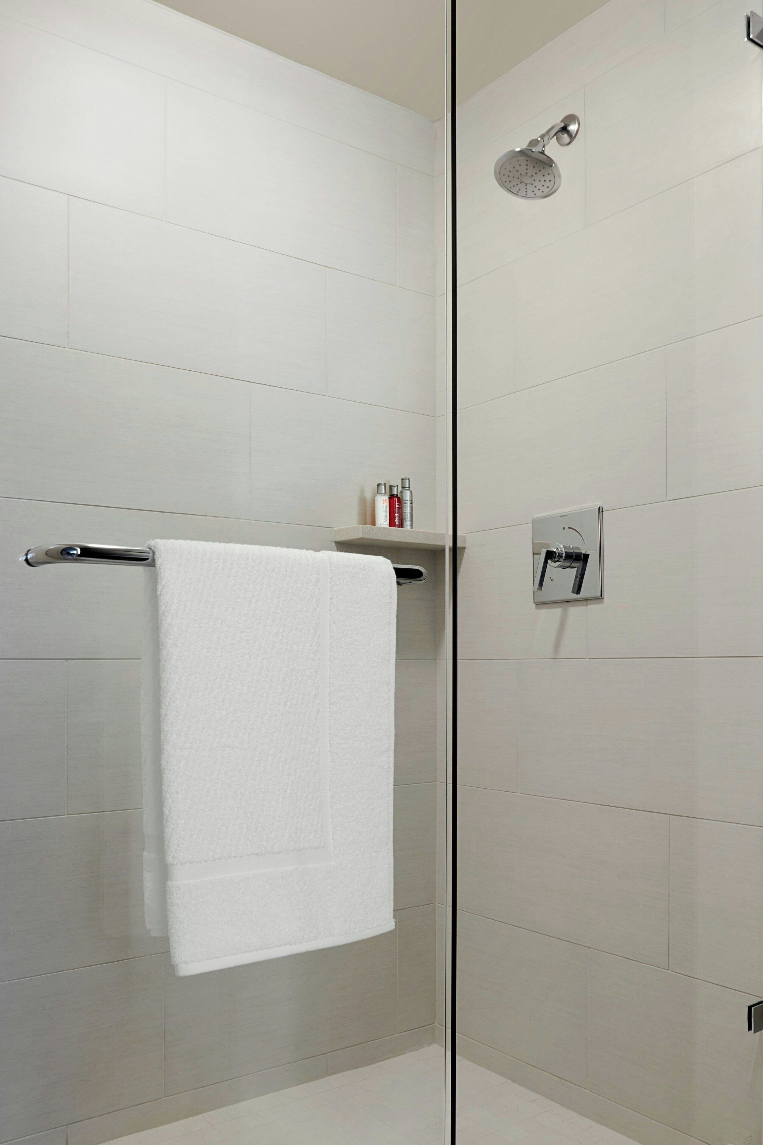 Un baño moderno con cabina de ducha de vidrio, una toalla blanca colgada en un perchero metálico y un estante pequeño con artículos de aseo.