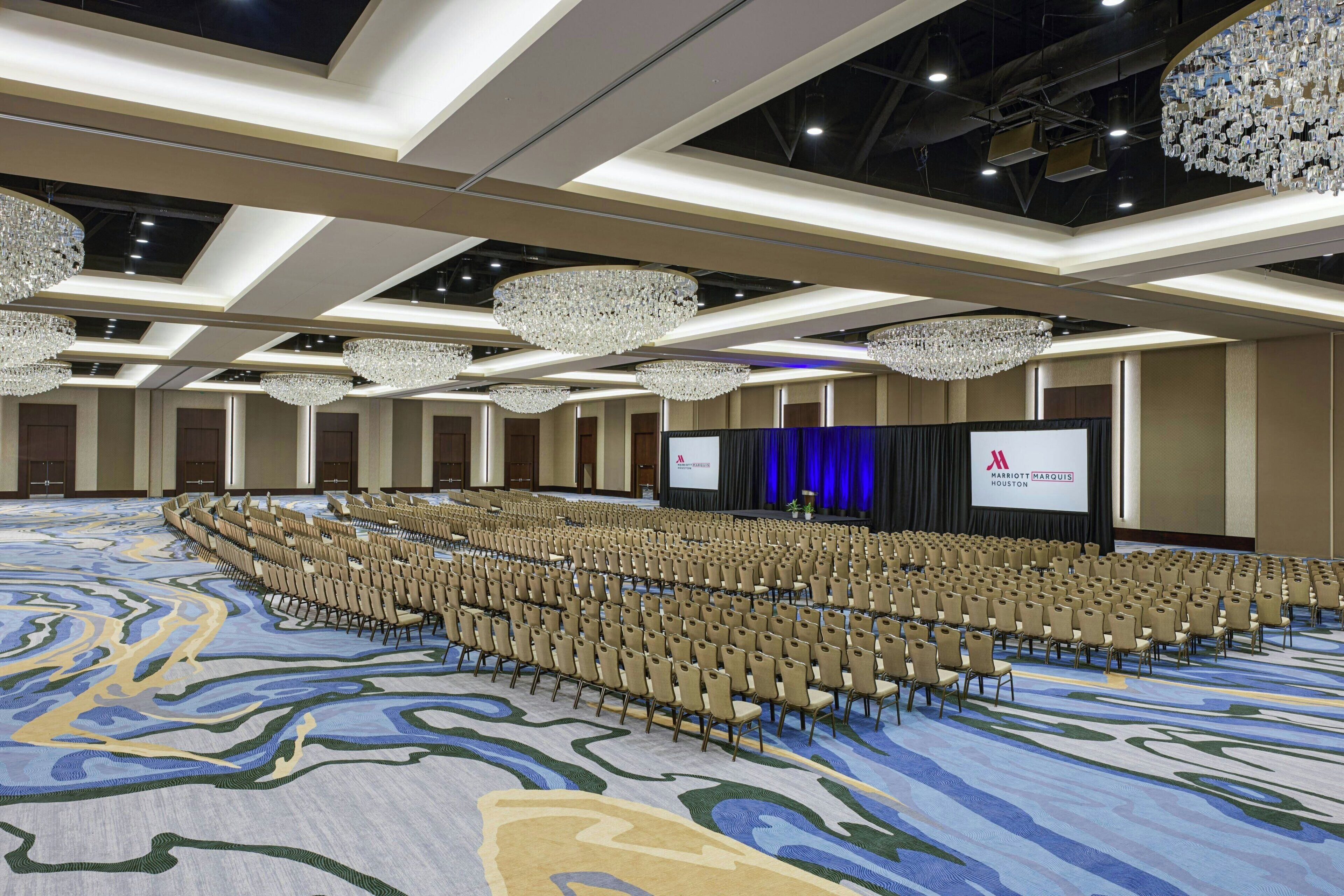 A large conference room with rows of chairs, a stage with a screen, and chandeliers.