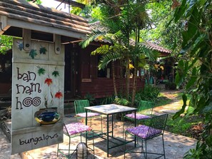 Ein Bungalow mit einer Betonstütze, auf der 'ban mam hoo' und 'Bungalows' steht. Grüne Metallstühle und ein Tisch mit violetten Kissen sind ebenfalls vorhanden.