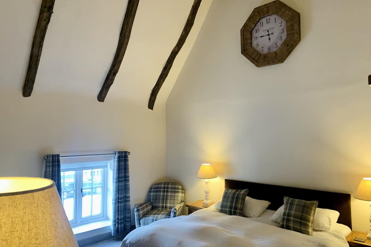 A hotel room with a bed, two bedside lamps, a plaid armchair, a window with curtains, and a wooden ceiling beam.