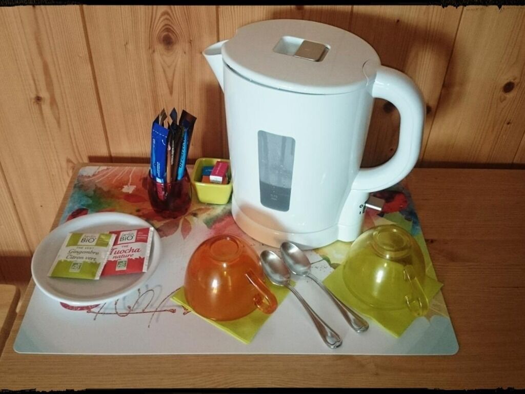 Een witte waterkoker, twee lepels, een gele en een oranje theepot, en een klein bord met theezakjes op een houten tafel.