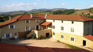 Un immeuble à plusieurs étages, doté d’une façade en pierre et de toits recouverts de tuiles rouges, situé dans un paysage rural avec des collines en arrière-plan.