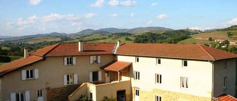 A multi-story building with a stone facade and red-tiled roofs, set in a rural landscape with hills in the background.