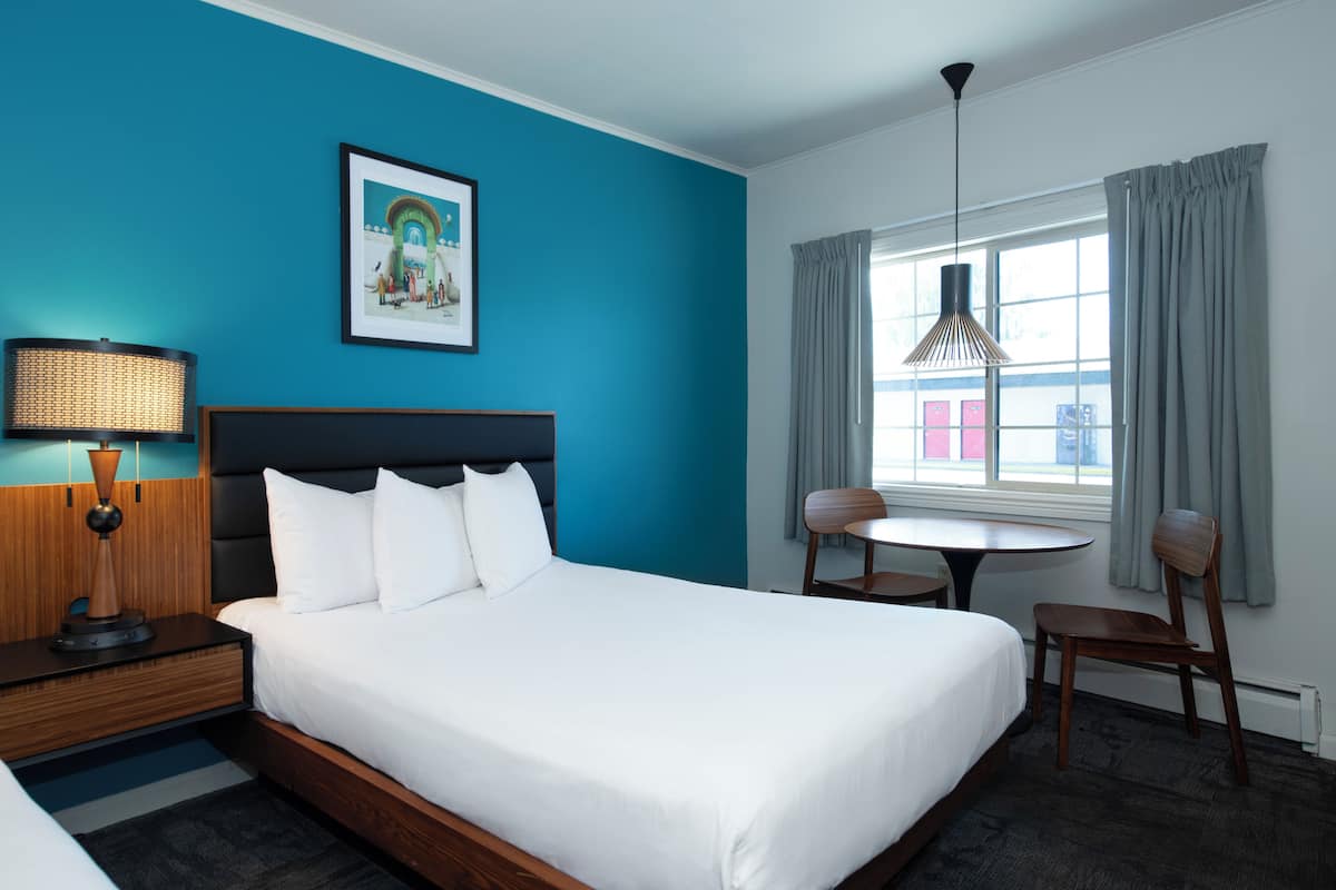 A hotel room with a large bed, a small table, two chairs, a lamp, and a window with curtains.
