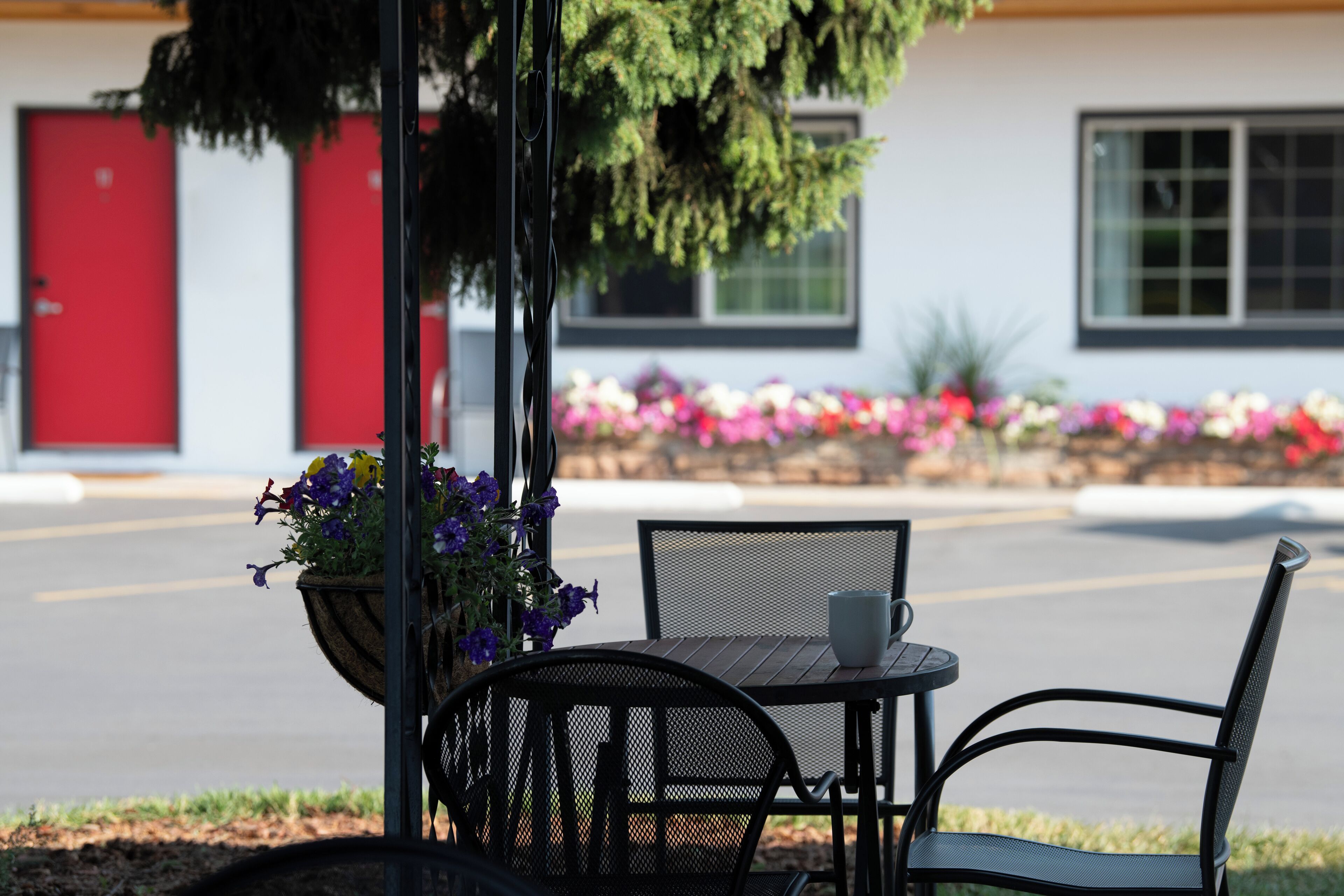 Un área de estar al aire libre con una mesa y sillas, una jardinera con flores, y un edificio con puerta roja y ventanas al fondo.