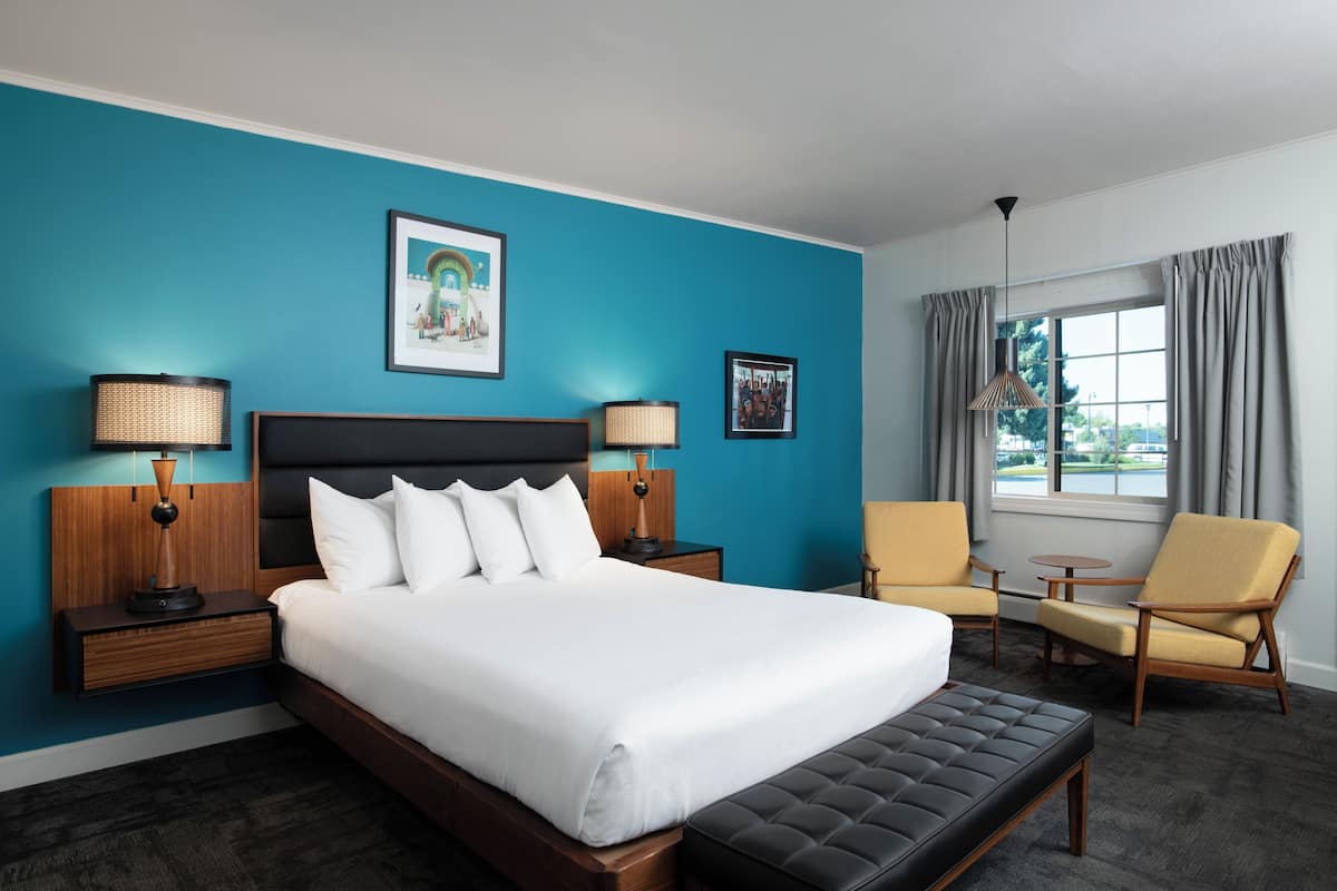 A hotel room with a large bed, two bedside tables with lamps, a chair, a small table, and a window with curtains.