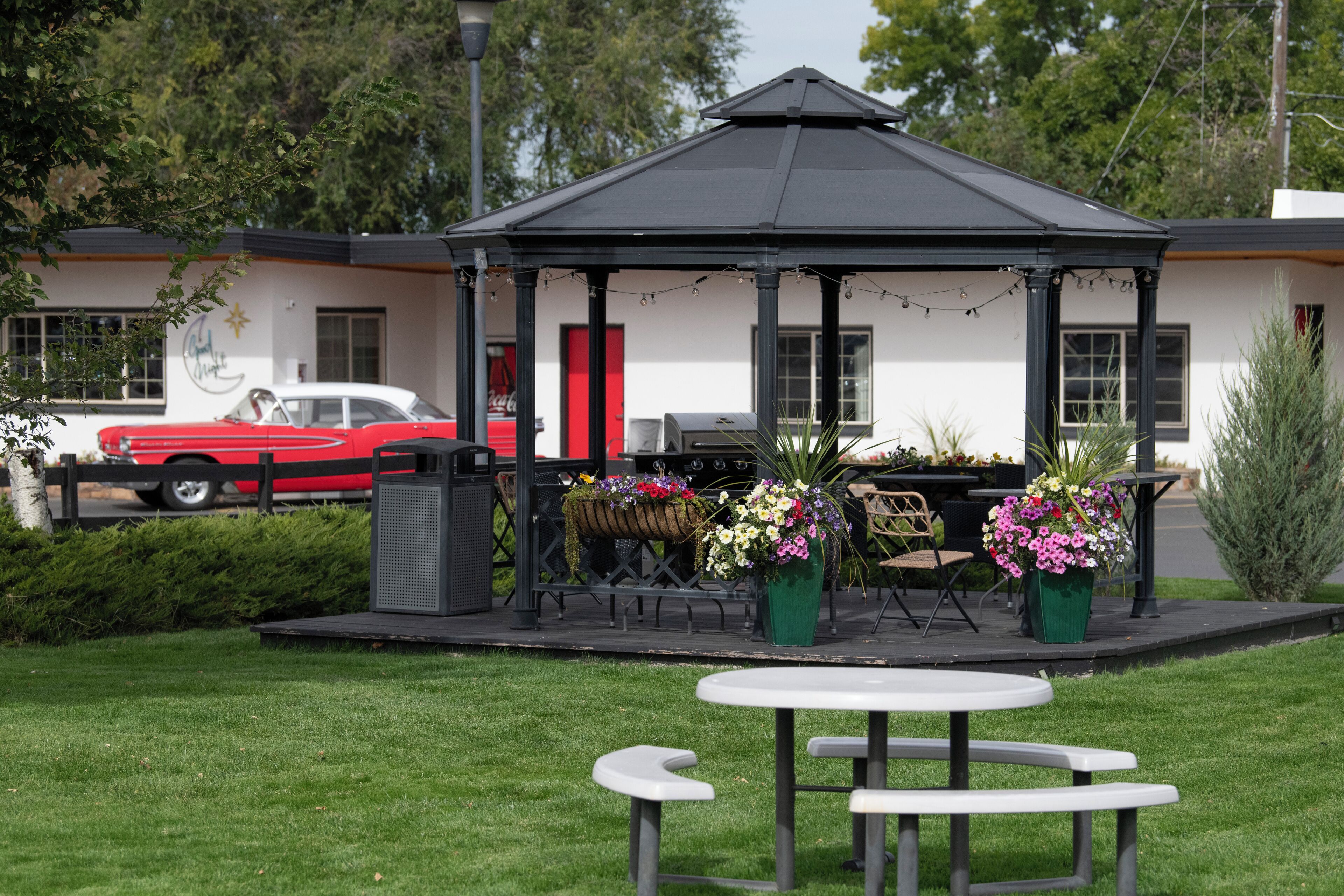 Un kiosque doté d’un toit noir et de murs blancs, entouré d’un jardin où sont disposées diverses plantes et fleurs en pot.