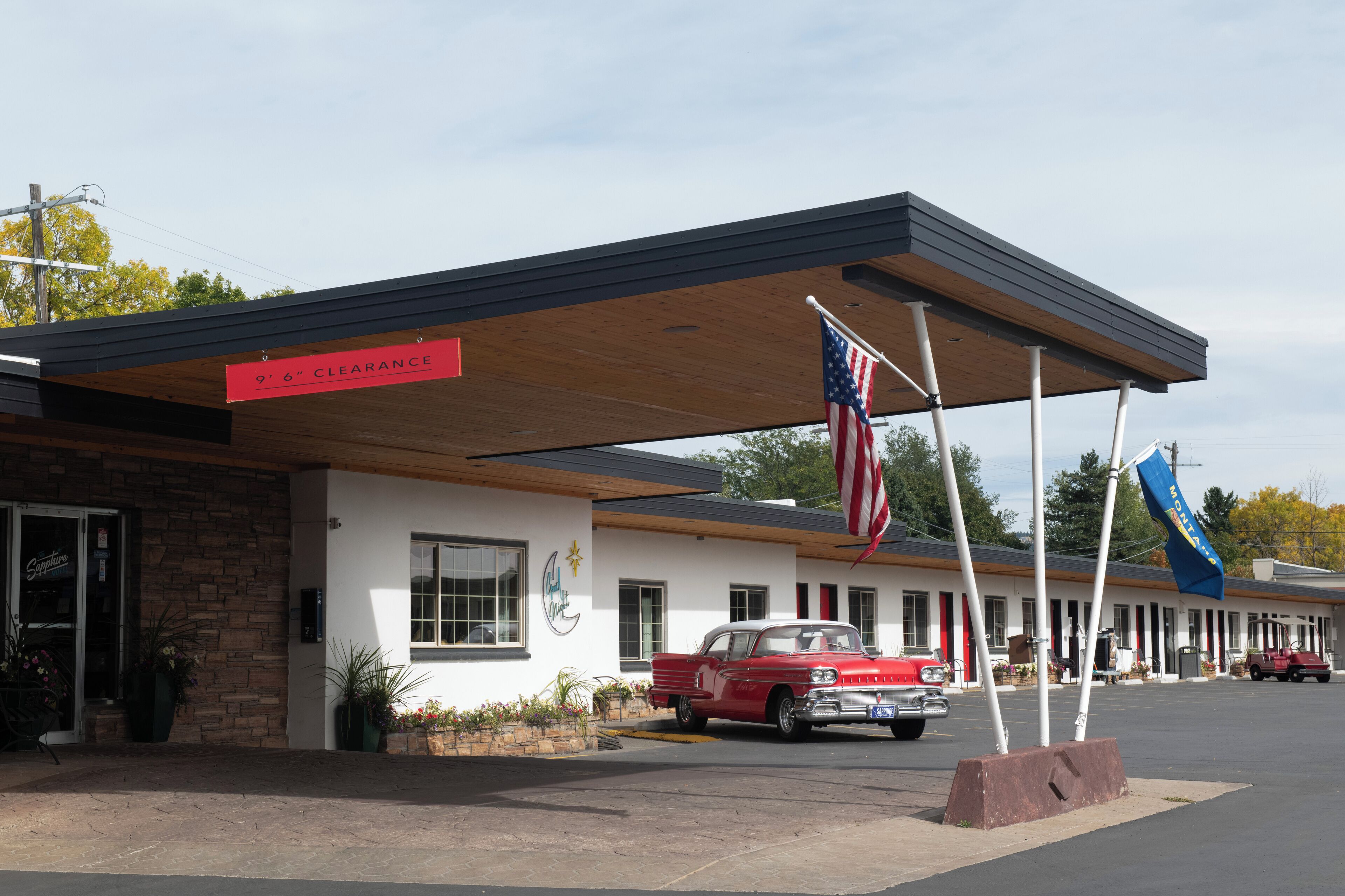 A motel with a 6" clearance sign, American flags, and a vintage red car parked in front.