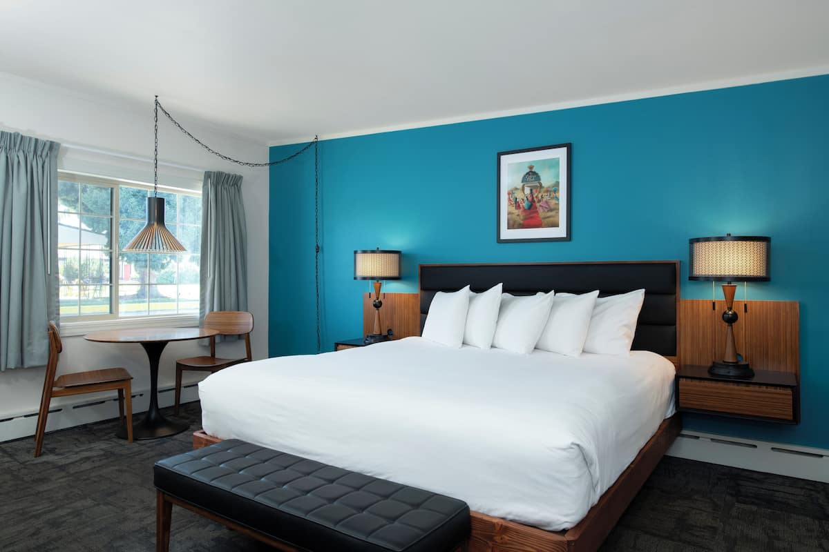 A hotel room with a large bed, two bedside tables with lamps, a small table and chair, and a window with curtains.